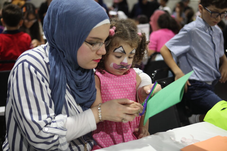A person wearing a hijab holding a child in her lap as they cut shapes out of paper. The child has face paint on.