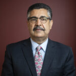 Picture of elderly man wearing a suit, tie, glasses and has a moustache and graying hair in front of a burgundy background.